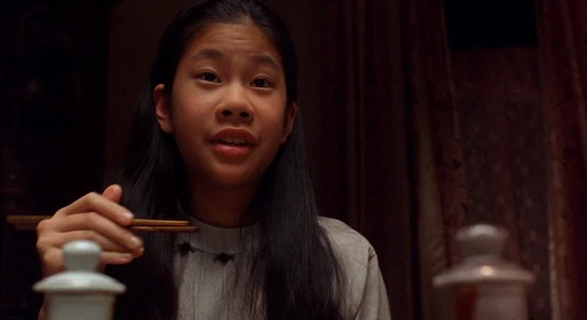 Double Happiness (1994) movie still: medium — A dimly lit room with dark brown curtains in the background. A young woman holds a pair of wooden…
