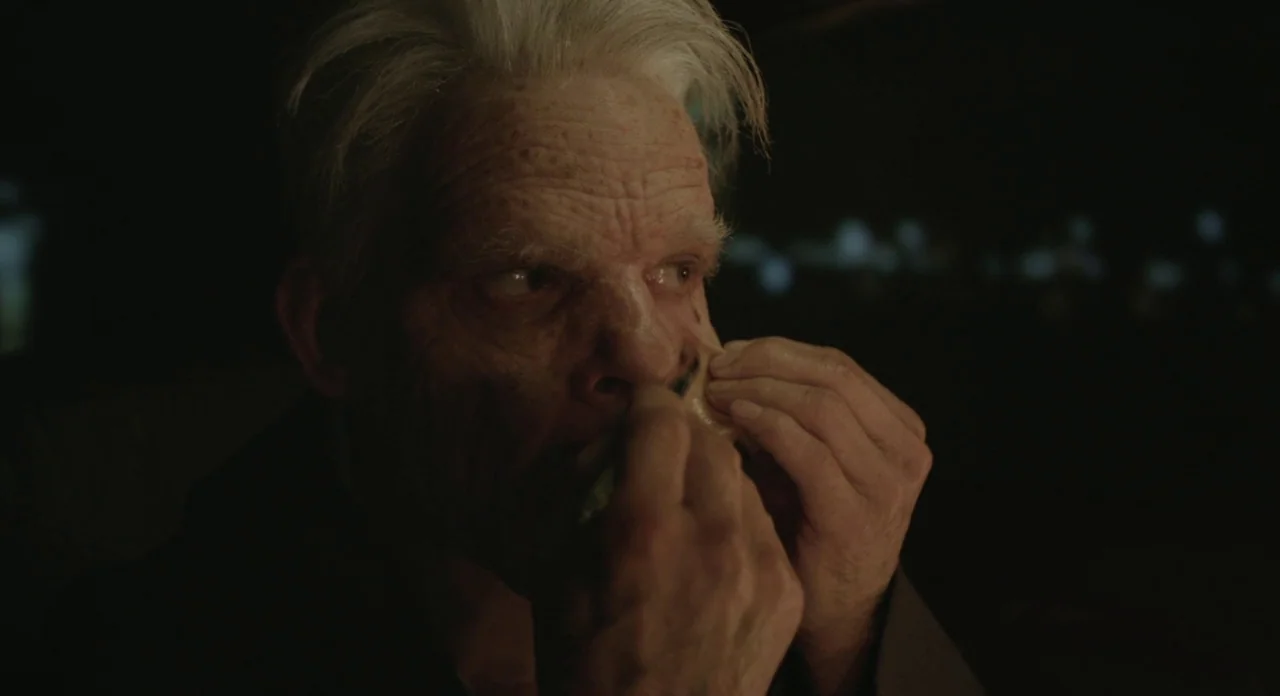 Holy Motors (2012) movie still: closeup — A dark, dimly lit environment provides the backdrop for the close-up shot. An elderly man with white hair…