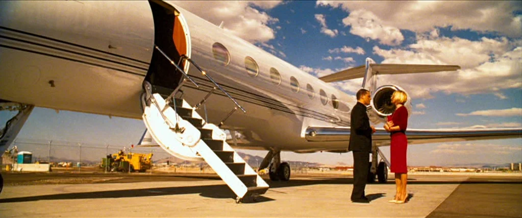 Ocean’s Thirteen (2007) movie still: wide — A private business jet is parked on a sunlit airfield tarmac with mountains visible. A man in a suit and…