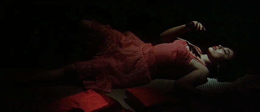 Jigoku (1960) movie still: medium — A dim, dark room features a straw-textured floor mat and deep red fabric elements. A woman in a red dress rests…