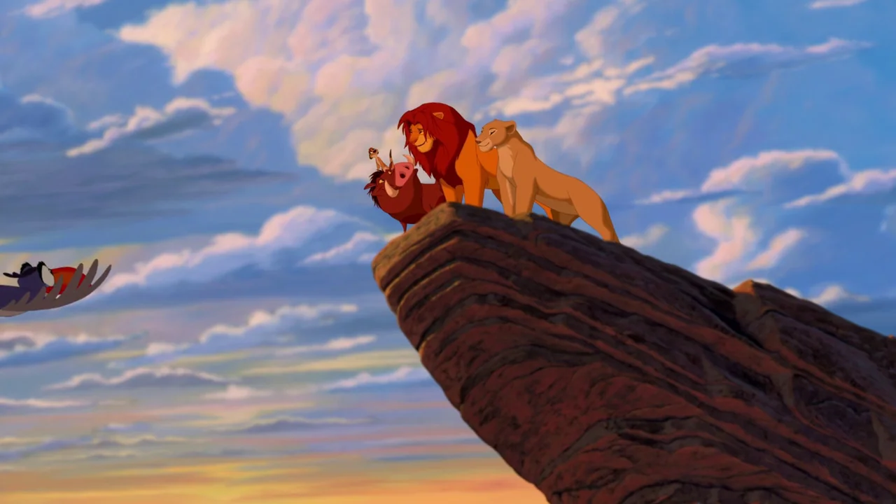 The Lion King (1994) movie still: wide — A high, jagged rock formation rises into a vibrant blue sky filled with clouds. Four animals stand together…