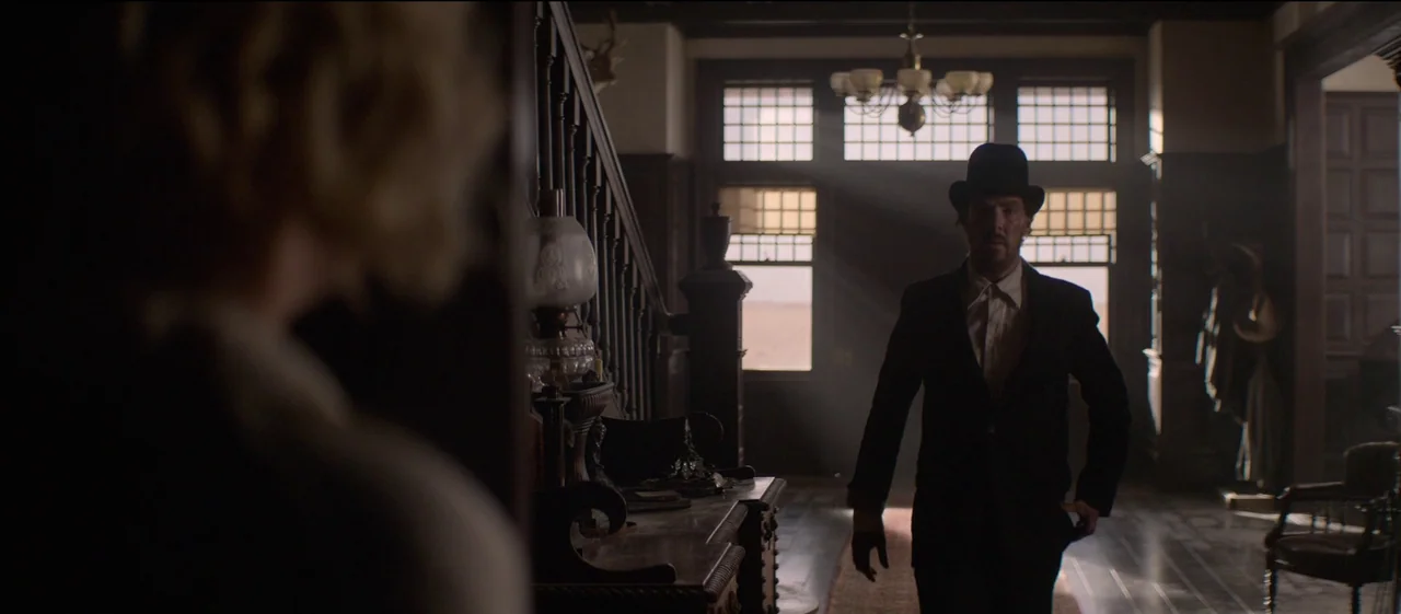 The Power of the Dog (2021) movie still: medium — A dimly lit grand hallway of a 1920s house features a dark wood staircase. A man wearing a top hat…