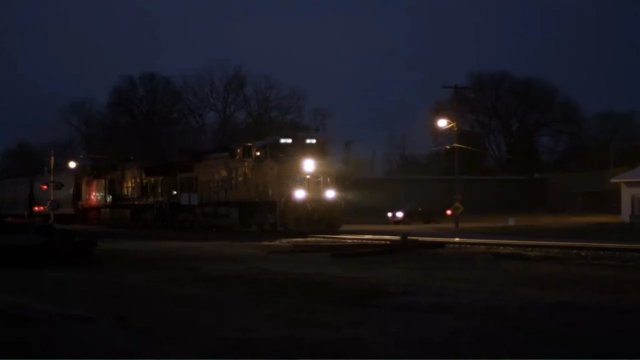 Rich Hill (2014) movie still: wide — A rural railroad crossing at night with a large locomotive train approaching the viewer. The front train lights…
