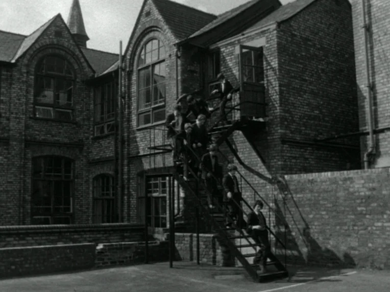 Children (1976) movie still: wide — A dark brick building features an external metal staircase leading to an upper floor. A group of schoolboys in…