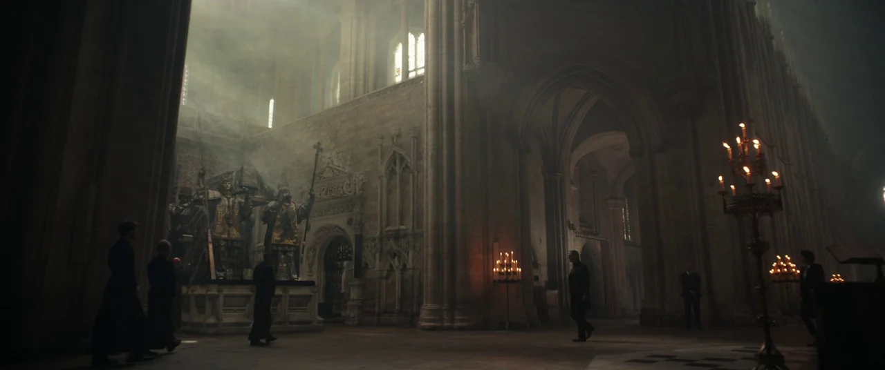 Assassin’s Creed (2016) movie still: wide — A grand stone cathedral nave features large decorative tombs and rows of floor candelabras. Several…