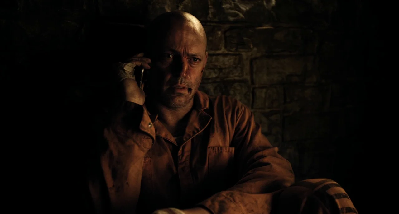 Brawl in Cell Block 99 (2017) movie still: closeup — A man sits against a rough-hewn stone wall in a dimly lit enclosure. He holds a smartphone to…