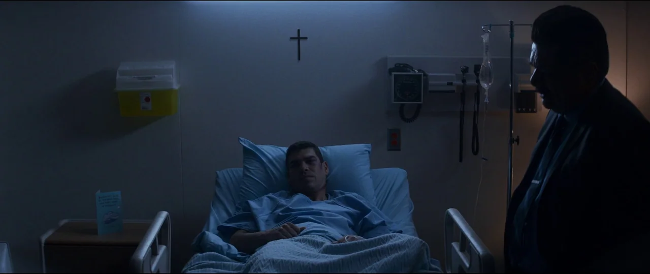 El Chicano (2018) movie still: medium — A dim hospital room features a blue-tinted environment with a small crucifix on the wall. An injured man…