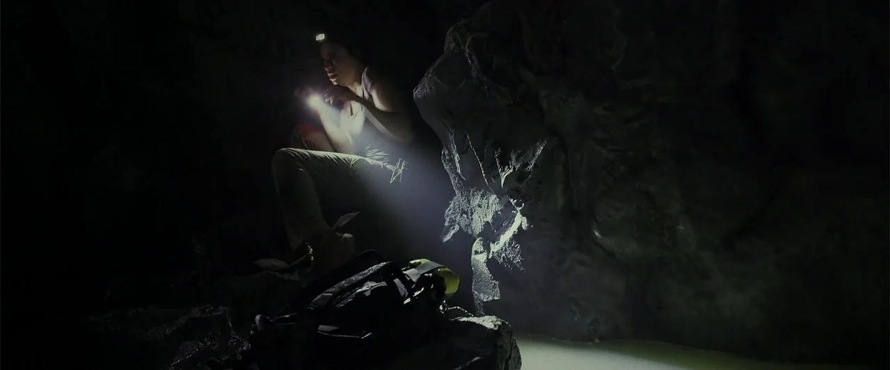 Black Water: Abyss (2020) movie still: medium — A dark, cramped interior of a rock cave with damp walls and pooling water. A woman in a tank top…