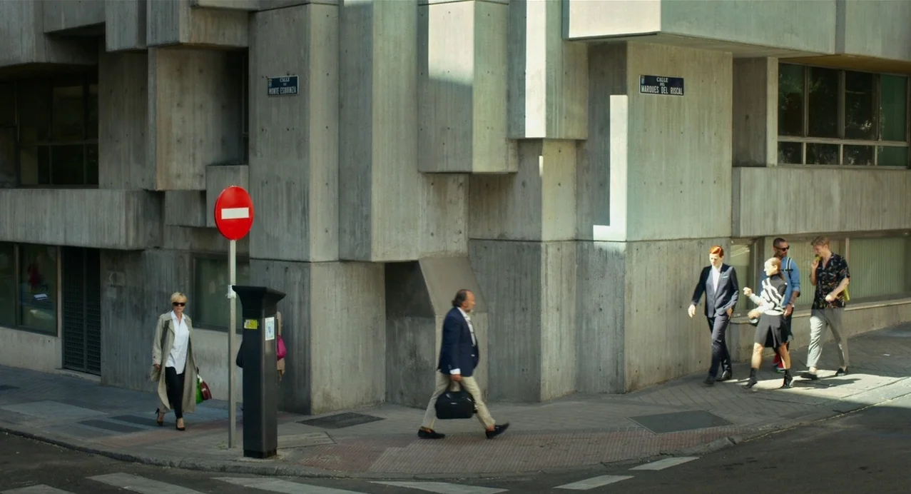 Julieta (2016) movie still: wide — A brutalist concrete office building occupies the corner of a city street intersection. Several pedestrians walk…