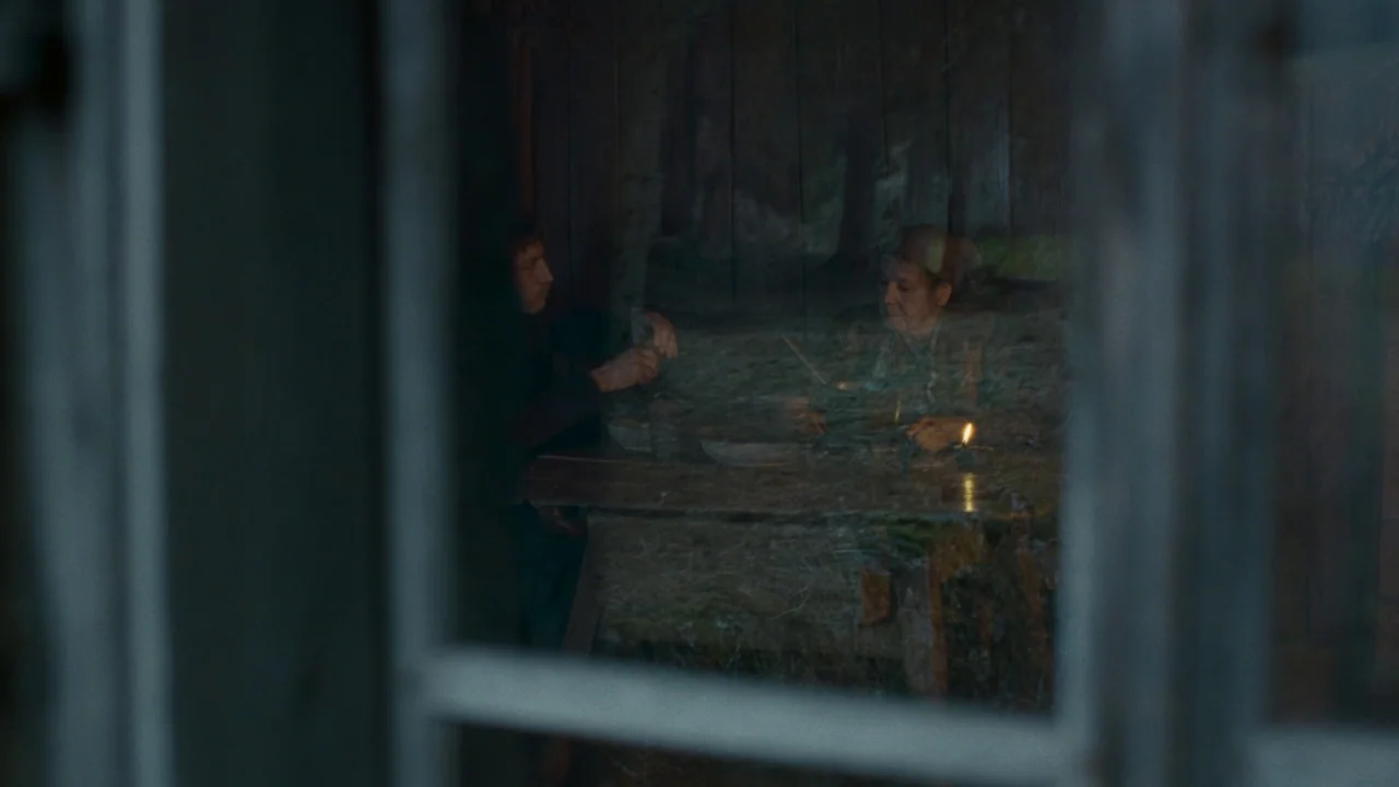 The Devil’s Bath (2024) movie still: medium — A rustic wooden interior is viewed through a window frame with strong reflections. Two people are…