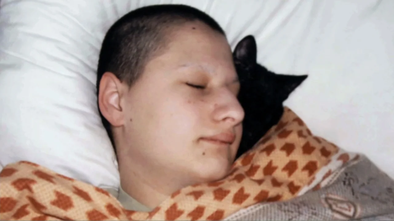 Mommy Dead and Dearest (2017) movie still: closeup — A person lies on a white pillow in a bed with a blanket. A black cat rests its head beside the…