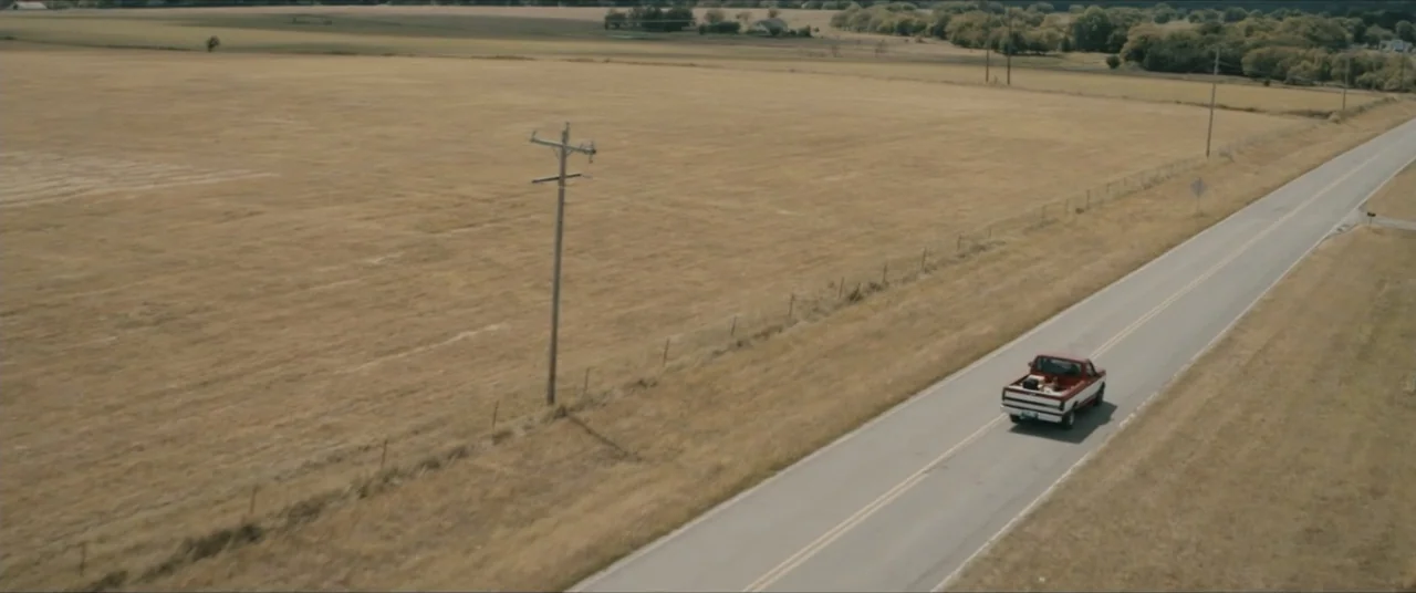 Possessions (2024) movie still: wide — A dry, grassy field borders an asphalt road stretching into the distance. A red and white pickup truck drives…