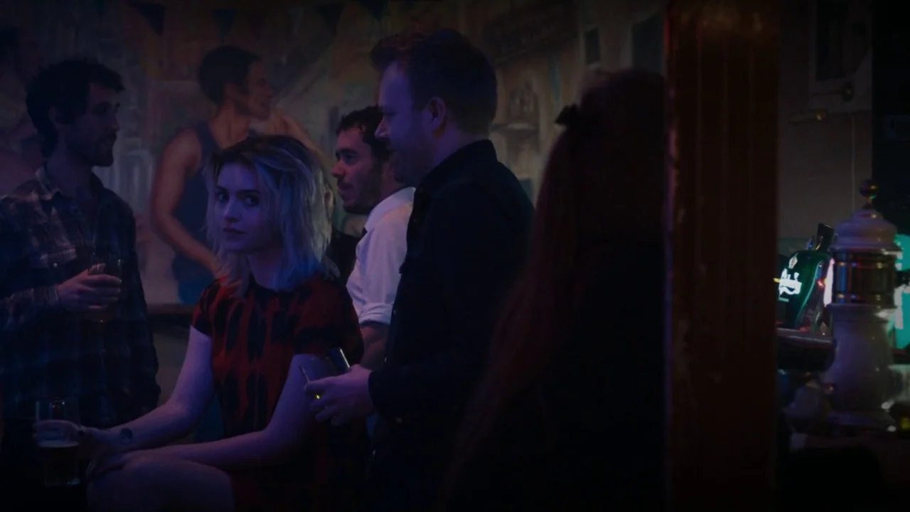 A Date for Mad Mary (2016) movie still: medium — A dimly lit pub with a wall mural and blurred patrons in the background. A young woman sits at a…