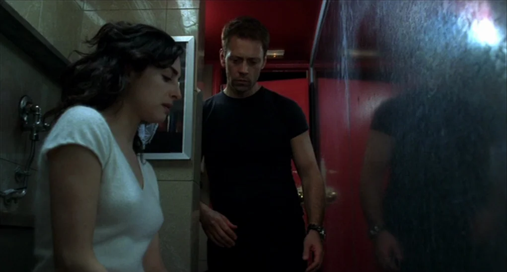 Anatomy of Hell (2004) movie still: medium — A dimly lit restroom with tiled walls features a bright red painted door frame. A man and a woman stand…