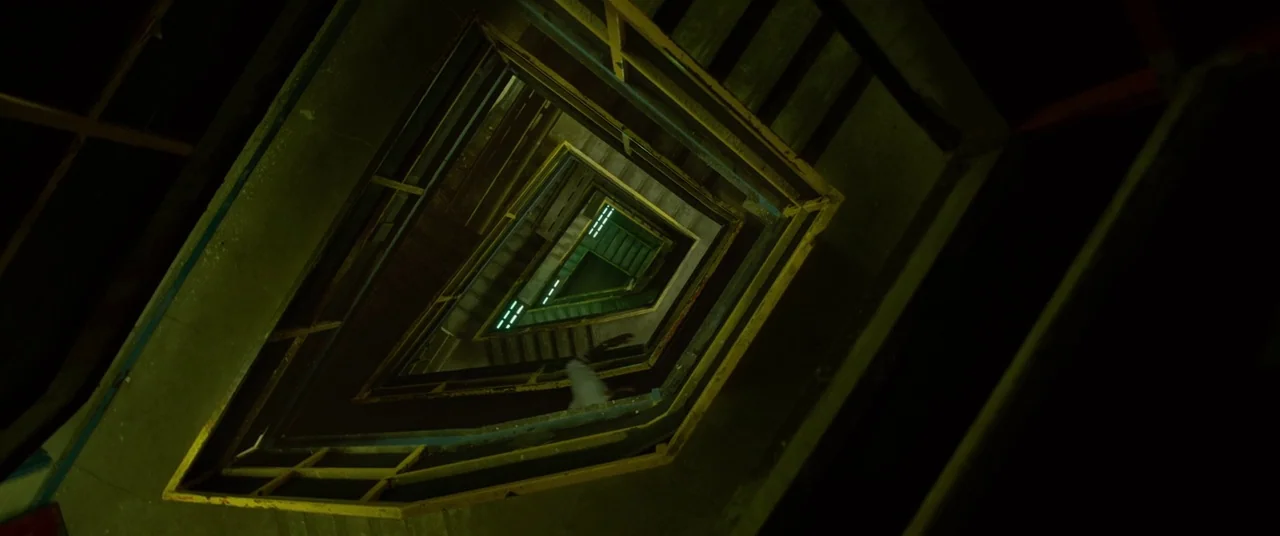 Spiral (2021) movie still: establishing — A dark, triangular stairwell features concrete steps and yellow metal railings. The perspective looks…