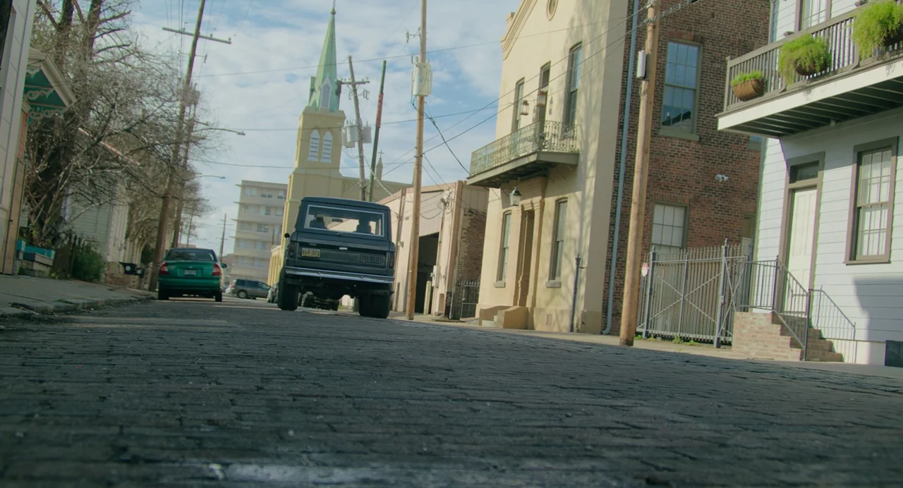 Deep Water (2022) movie still: wide — A historic residential street with cobblestones, old brick buildings, and a church spire background. A dark…