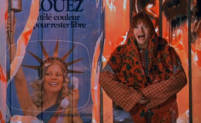 Sweet Movie (1974) movie still: medium — A person stands in front of a colorful, torn advertisement for a television service. The ad features an…