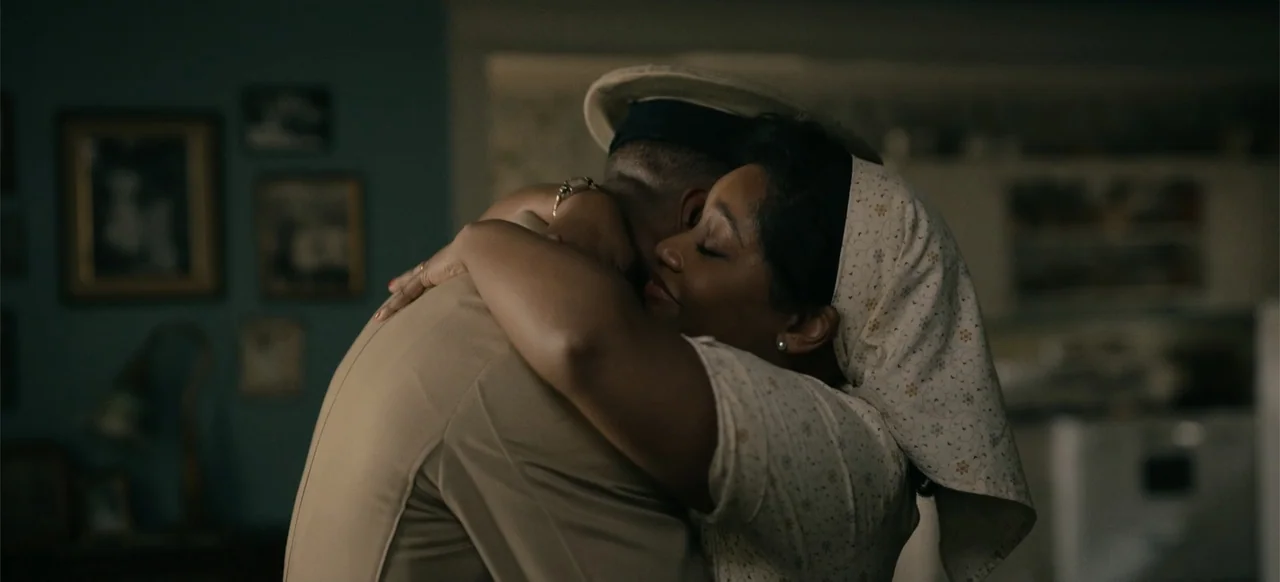 Devotion (2022) movie still: medium — An interior living room with framed pictures on a teal-colored wall. A woman in a floral headscarf embraces a…