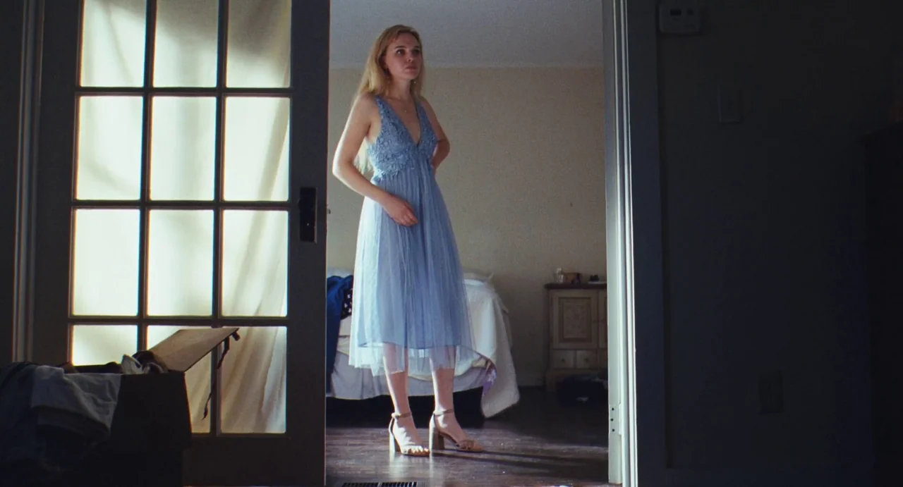 The Giant (2019) movie still: fullbody — A woman stands in a wooden doorway connecting a bright hallway to a dimmer room. The room beyond features a…