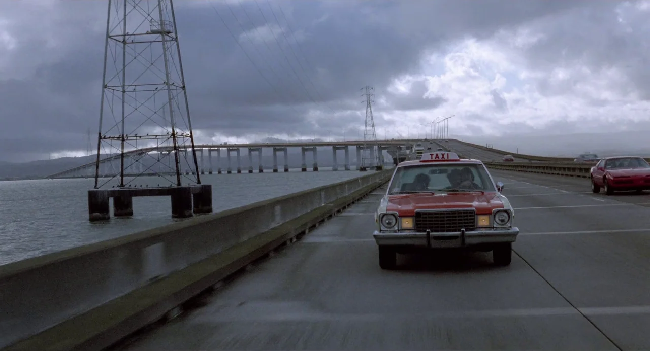 Sid and Nancy (1986) movie still: wide — A long concrete bridge spans over a grey body of water under overcast skies. A vintage taxi drives along…