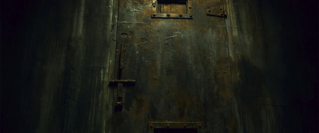 The Unholy (2022) movie still: medium — A heavy rusted metal door features a small observation slot and a sliding bolt lock. The dark, weathered…