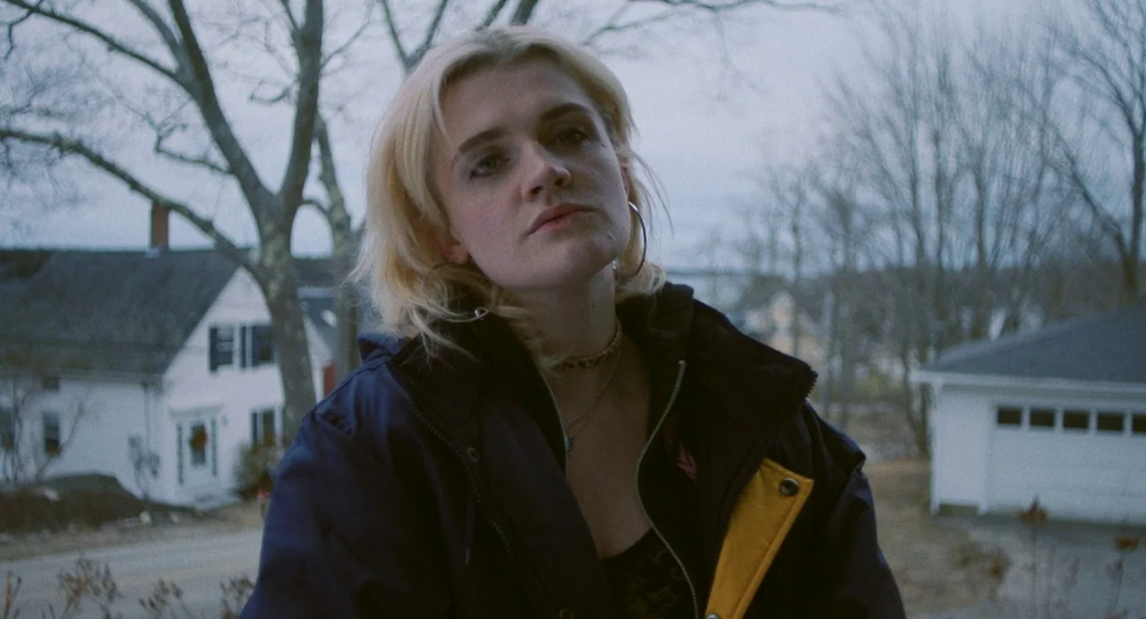 Blow The Man Down (2019) movie still: medium — A residential neighborhood with bare-limbed trees and white houses in the background. A young woman…
