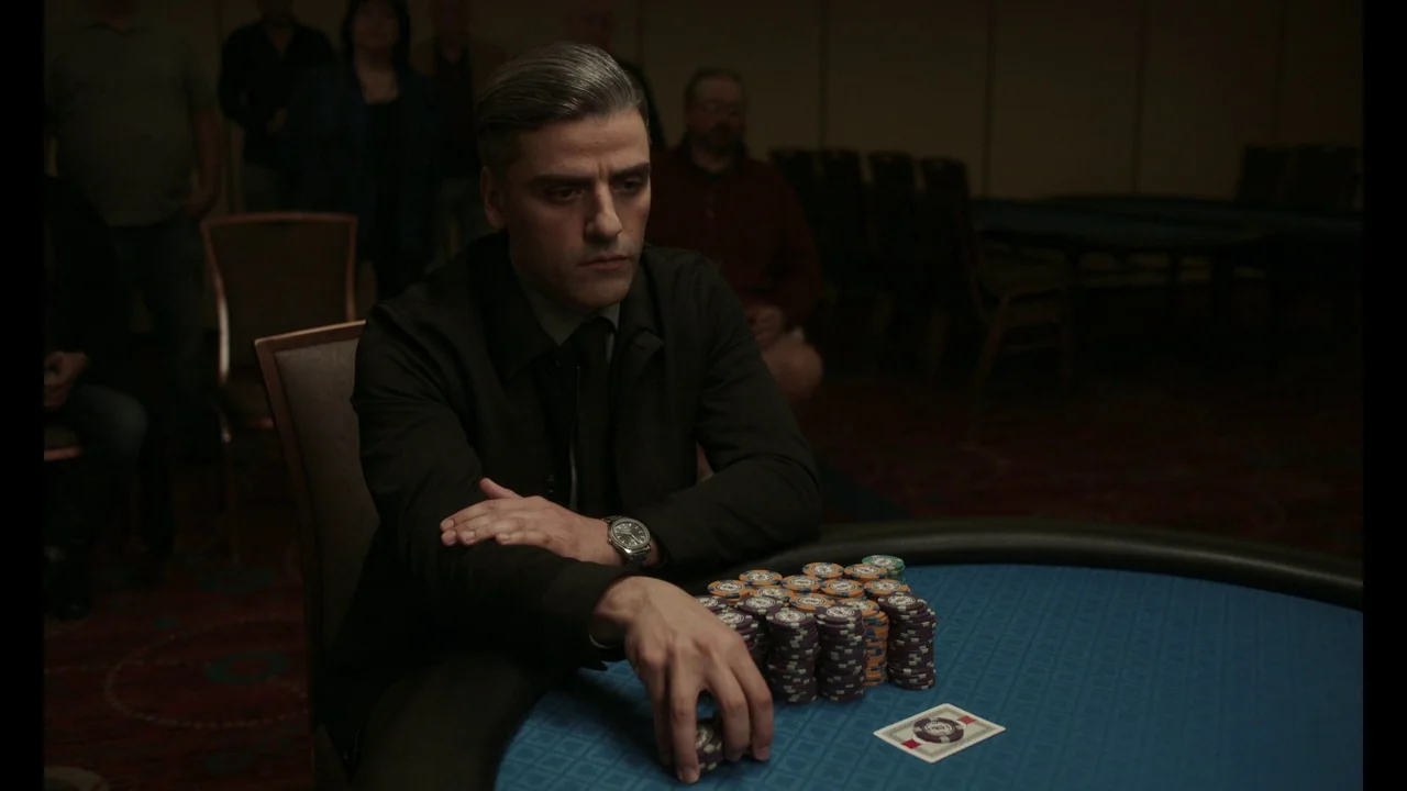 The Card Counter (2021) movie still: medium — A dimly lit room contains rows of tables and scattered figures in the background. A man sits at a blue…