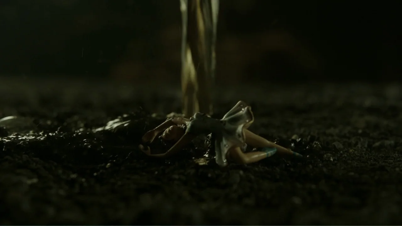 A Cure for Wellness (2017) movie still: closeup — A small plastic doll lies on a dark, wet gravel surface in the dark. A thin stream of liquid falls…