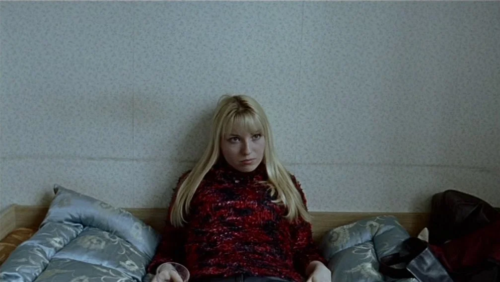 Import Export (2007) movie still: medium — A small room with light grey patterned wallpaper features a bed with blue satin-like pillows. A young…