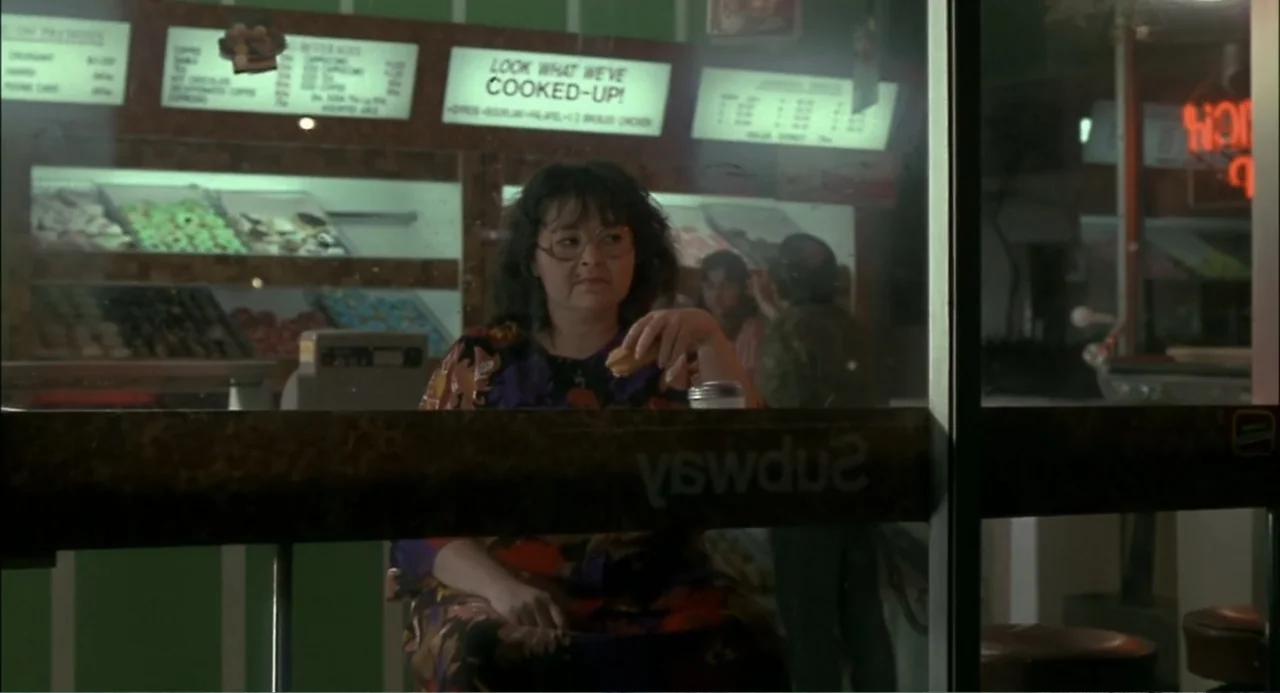 She-Devil (1989) movie still: medium — A woman sits behind a glass partition inside a fast-food eatery at night. She holds a small food item while…