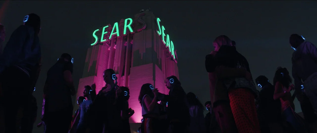 The Greatest Hits (2024) movie still: wide — A crowd gathers on a dark rooftop under a large glowing Sears neon sign. Young people wearing glowing…