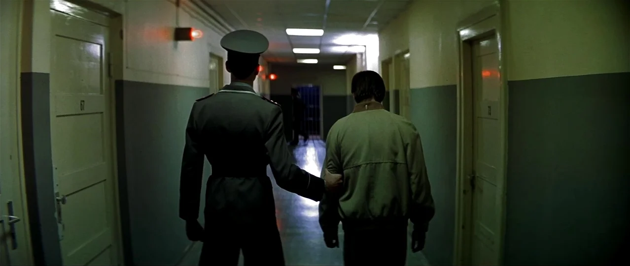 The Lives of Others (2006) movie still: fullbody — A long, dimly lit institutional corridor features grey walls and numbered wooden doors on either…