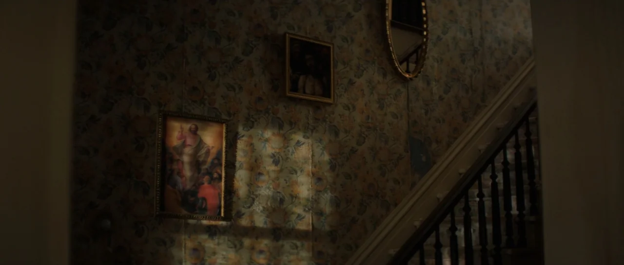 The Front Room (2024) movie still: establishing — An interior staircase features floral wallpaper and two framed paintings hung on the wall. The…