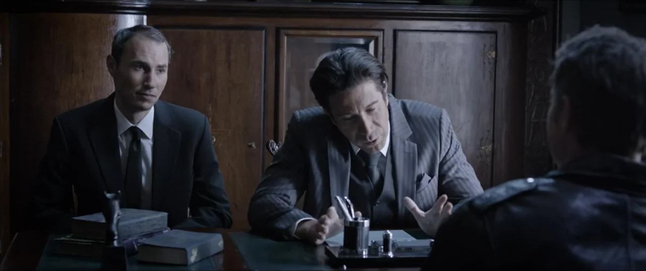 Room 6 (2006) movie still: medium — A dimly lit room with dark wood paneling and a leather-topped desk. Two men in formal business suits talk to…