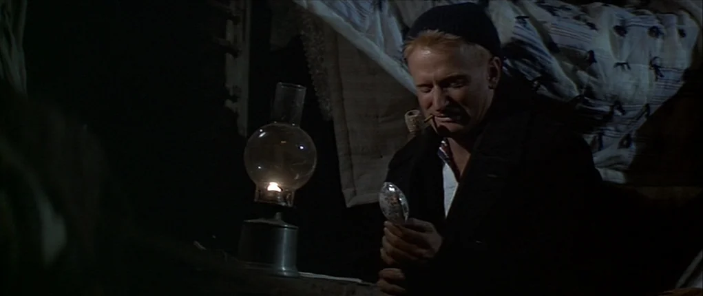 Popeye (1980) movie still: medium — A dimly lit cabin interior features wooden surfaces and patterned fabric bedding in the background. A man sits…