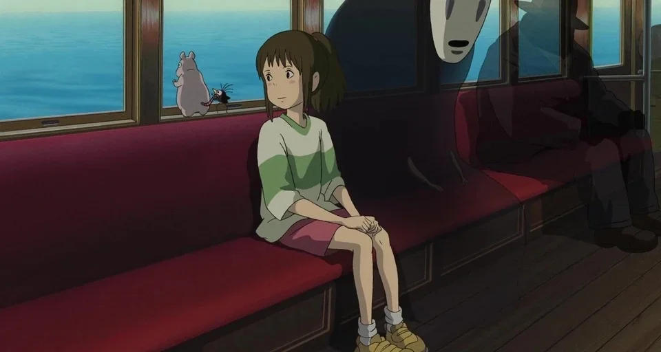 Spirited Away (2001) movie still: medium — A quiet, wood-paneled train interior moves over a calm, bright blue body of water. A young girl sits on a…