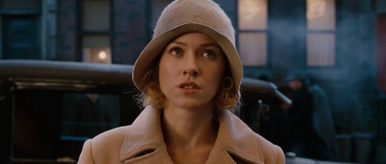 King Kong (2005) movie still: closeup — An urban setting with blurred buildings and vintage vehicles in the background. A woman in a tan coat and…