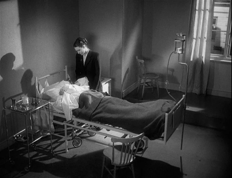 Music in Darkness (1948) movie still: wide — A stark room features a metal bed frame, medical cart, and wooden chairs. A woman in a dark suit leans…