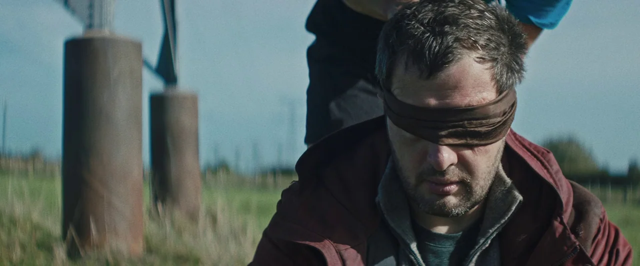 Vincent Must Die (2023) movie still: closeup — A man kneels in an open field near two cylindrical rusted metal pillars. He wears a brown blindfold…