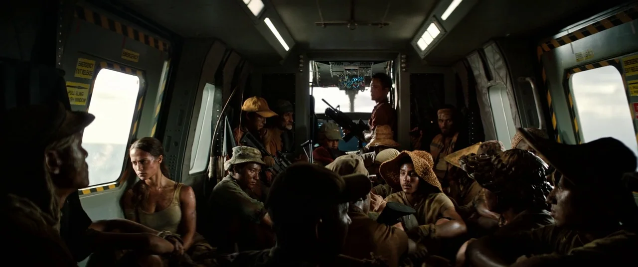 Tomb Raider (2018) movie still: wide — The interior of a cramped cargo helicopter contains many passengers seated in rows. People in casual work…