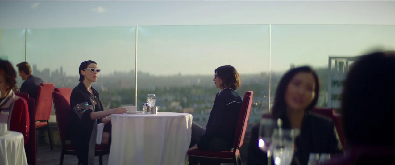 The Nowhere Inn (2020) movie still: wide — A rooftop restaurant overlooks a distant cityscape during the late afternoon sun. Two women sit at a…