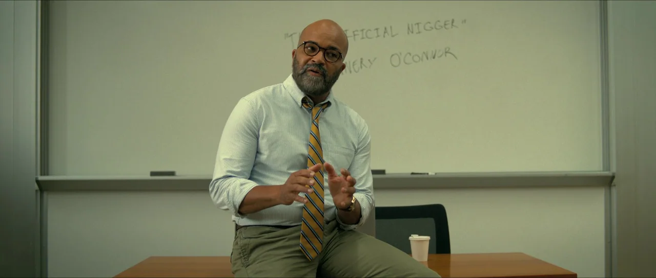 American Fiction (2023) movie still: medium — A professor sits on the edge of a desk in a classroom. Behind him a whiteboard displays partially…