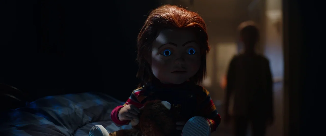Child's Play (2019) movie still: closeup — A dark bedroom at night contains a plastic doll sitting on a bed. The doll stares directly forward while…