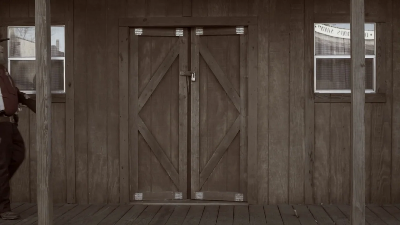 The Dalton Gang (2020) movie still: medium — A wooden building facade features a central locked double door flanked by two windows. A man in western…
