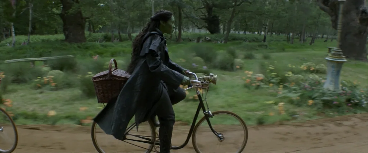 Wicked (2024) movie still: fullbody — A lush, overgrown meadow with dense trees features a worn dirt path. A woman with green skin rides a bicycle…