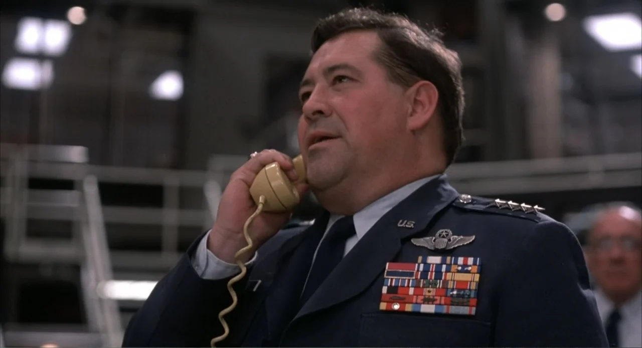 WarGames (1983) movie still: medium — A military command room features industrial metal structures and blurred technical equipment in background. A…