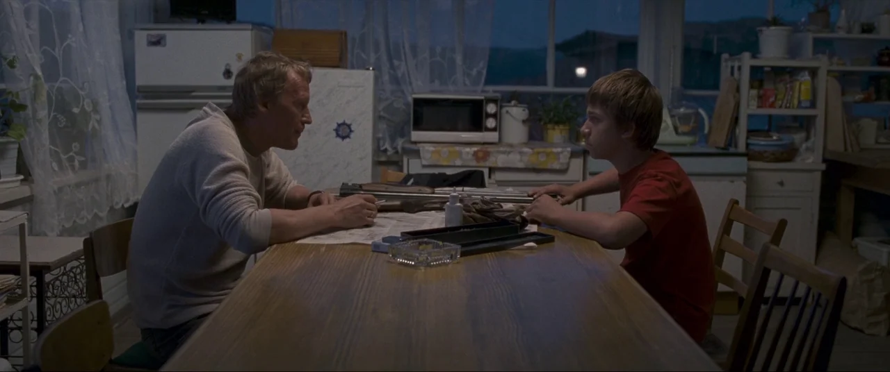 Leviathan (2014) movie still: medium — A modest dining room features a long wooden table cluttered with disassembled metal firearm parts. An adult…