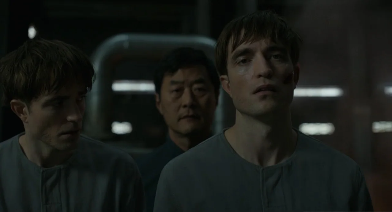 Mickey 17 (2025) movie still: medium — A sterile, dimly lit industrial laboratory contains dark walls and metallic background structures. Two…