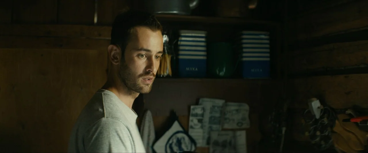 Invasion (2020) movie still: medium — A dark wooden rustic space features shelves lined with striped storage tins and papers. A young man turns his…