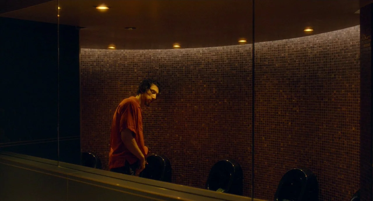Annette (2021) movie still: medium — A dimly lit restroom features a large curved wall covered in small brown tiles. A man stands near the wall in…